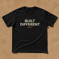 Built Different tee in black, printed with BUILT DIFFERENT in cream and quietly. in coral, laid flat on a tan background