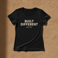 Built Different fitted tee in black, printed with BUILT DIFFERENT in cream and quietly. in coral, laid flat on a tan background