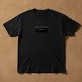 Debuggin' tee in black, printed with a small terminal window reading $ debuggin', laid flat on a tan background