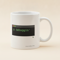 Debuggin' mug in white, printed with a small terminal window reading $ debuggin' in green