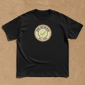 Pre-Revenue tee in black with a round PRE-REVENUE est. 2026 badge and green check mark, laid flat on a tan background