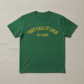 Est. Grind Varsity fitted tee in green, printed with THEY CALL IT LUCK EST. GRIND in gold varsity lettering, laid flat