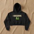 I Deserve AI cropped hoodie in black, printed with I DESERVE in cream and A.I. in green, laid flat on a tan background